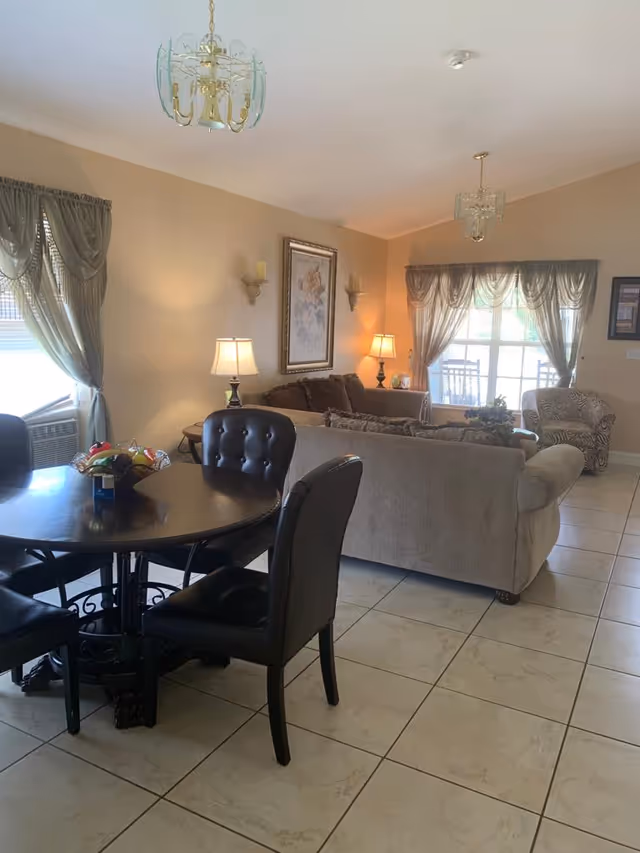A cozy living room and dining area with beige walls and tiled floor. The dining area features a round dark wooden table with four black cushioned chairs. The living room has a beige sofa, a patterned armchair, two table lamps, wall sconces, framed artwork, and windows with sheer curtains allowing natural light to enter.