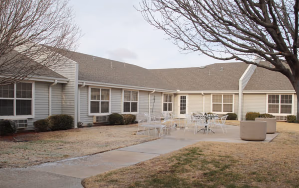 Outdoor courtyard area of a senior living facility with beige siding buildings surrounding a concrete patio. The patio has white metal tables and chairs. There are leafless trees and some shrubs around the building, with a grass lawn that appears dry.