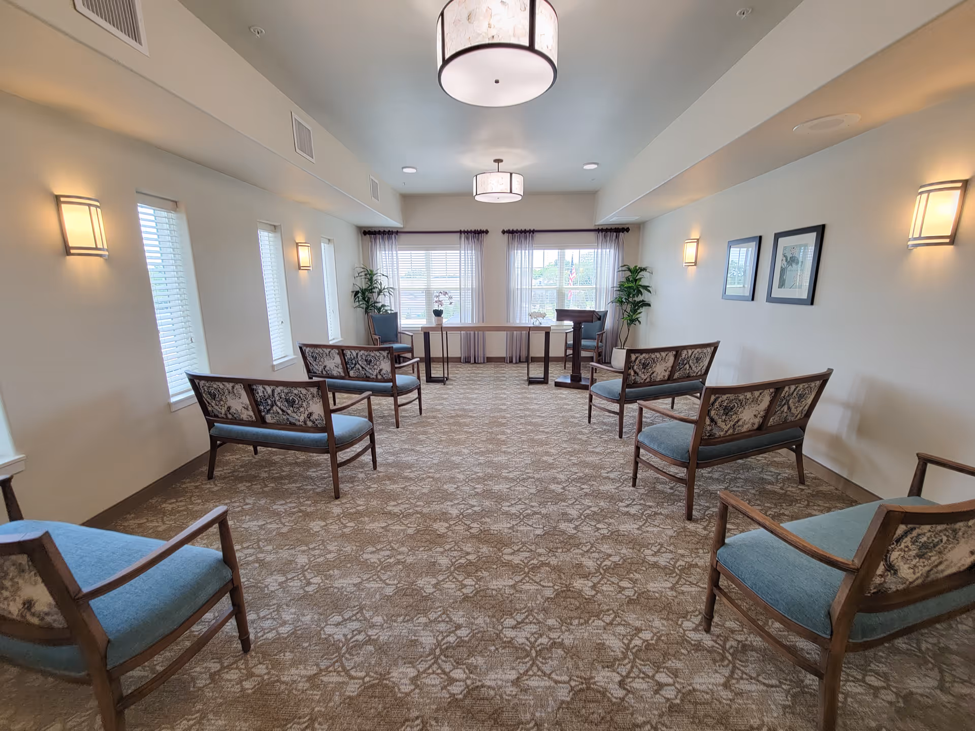 A well-lit room with patterned carpet and multiple wooden chairs with blue cushions arranged in two rows facing a wooden table near windows with sheer curtains. The walls are light-colored with wall sconces and framed pictures, and there are two ceiling lights and potted plants near the windows.