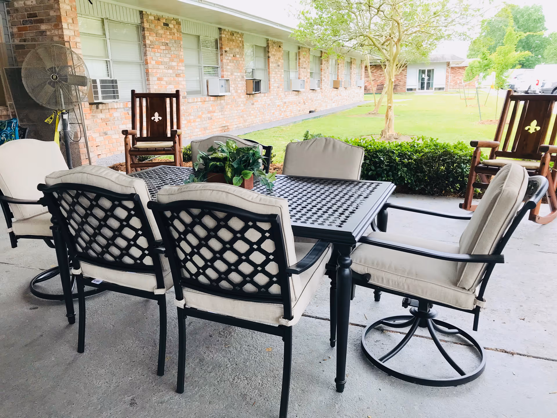 Patio dining set with cushioned metal chairs and a table on a covered concrete porch outside a brick senior living building.