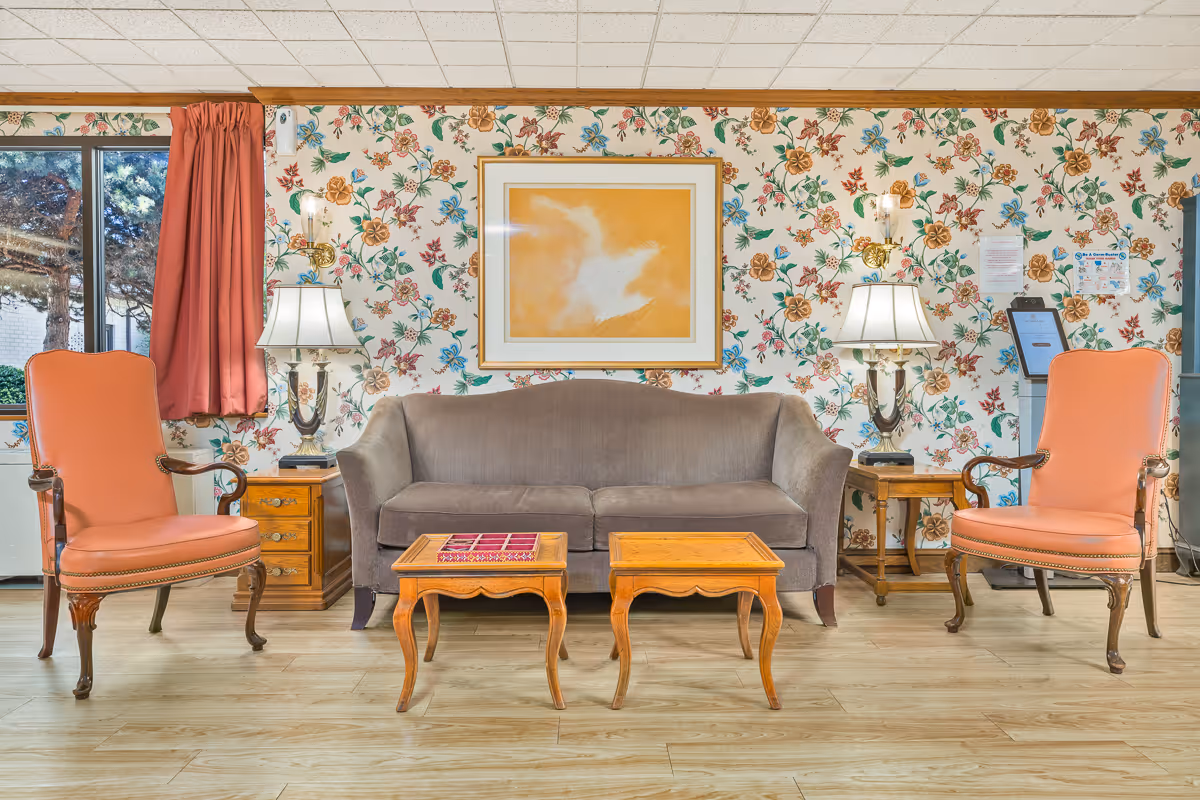 A floral-walled lounge with a gray sofa flanked by two coral armchairs, wooden coffee tables, lamps, and framed artwork.