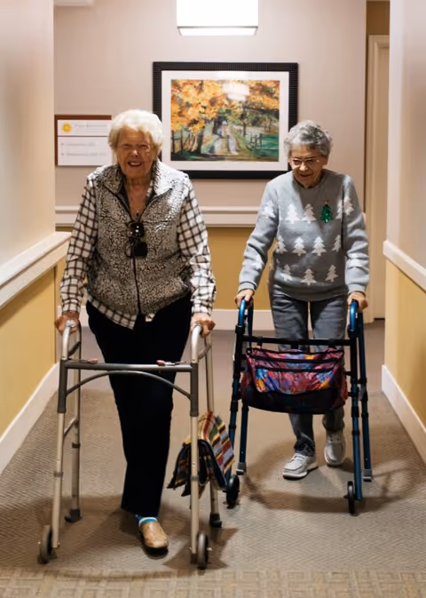 Two elderly women walking down a hallway using walkers. One woman is wearing a plaid shirt with a vest and sunglasses hanging from her neck, while the other is wearing a gray sweater with white tree patterns. There is a framed painting of an autumn scene on the wall behind them.