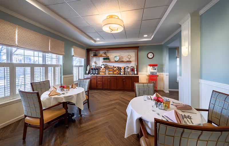 A bright dining room with round tables set for meals, upholstered chairs, large windows with shutters, and a buffet area with coffee and snacks.