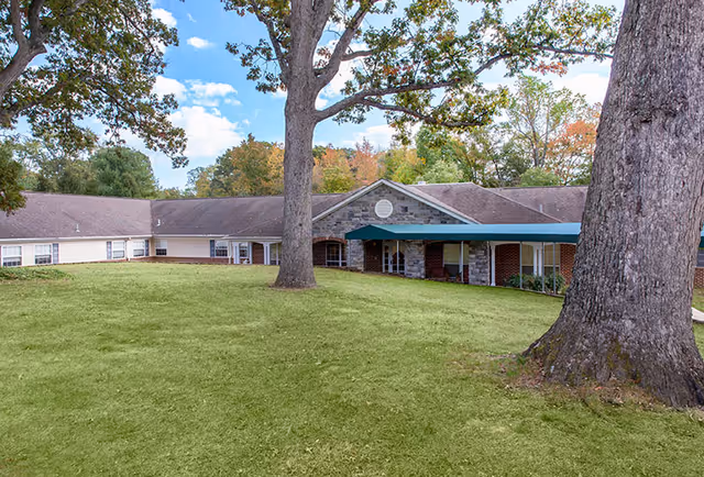 Single-story senior living facility exterior with a grassy front lawn, large trees, and a covered entrance.