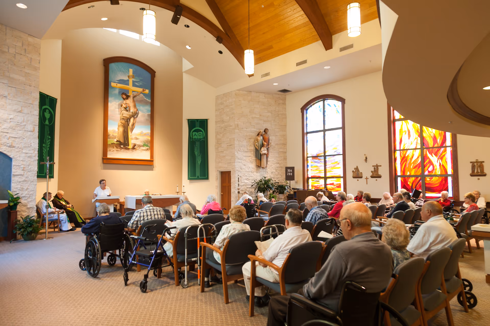 A group of elderly people seated in a chapel or worship room with stained glass windows and religious artwork, including a large painting of Jesus on the cross and statues. A woman stands at a podium speaking, while others listen attentively.