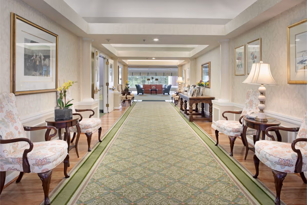 A well-lit hallway in a senior living facility with a long green patterned carpet running down the center. The walls are decorated with framed artwork and mirrors. On both sides of the hallway, there are floral upholstered chairs paired with small wooden tables, some holding lamps and flower arrangements. At the far end, a seating area with a red sofa and two blue armchairs is visible near large windows.
