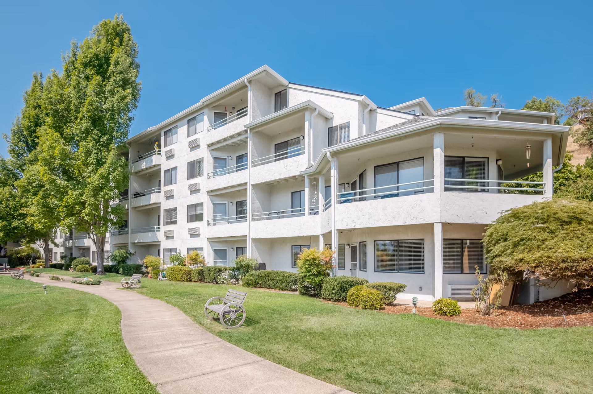 Exterior view of a multi-story white residential building with balconies, surrounded by green grass, bushes, and tall trees under a clear blue sky. A curved concrete pathway with benches runs along the front of the building.