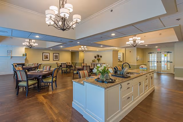 A spacious dining area in a senior living facility with multiple round tables and striped cushioned chairs. In the foreground, there is a large kitchen island with a granite countertop, a sink, and a flower arrangement. The room has warm lighting with chandeliers and recessed ceiling lights, wooden flooring, and light-colored walls. Double glass doors are visible in the background.