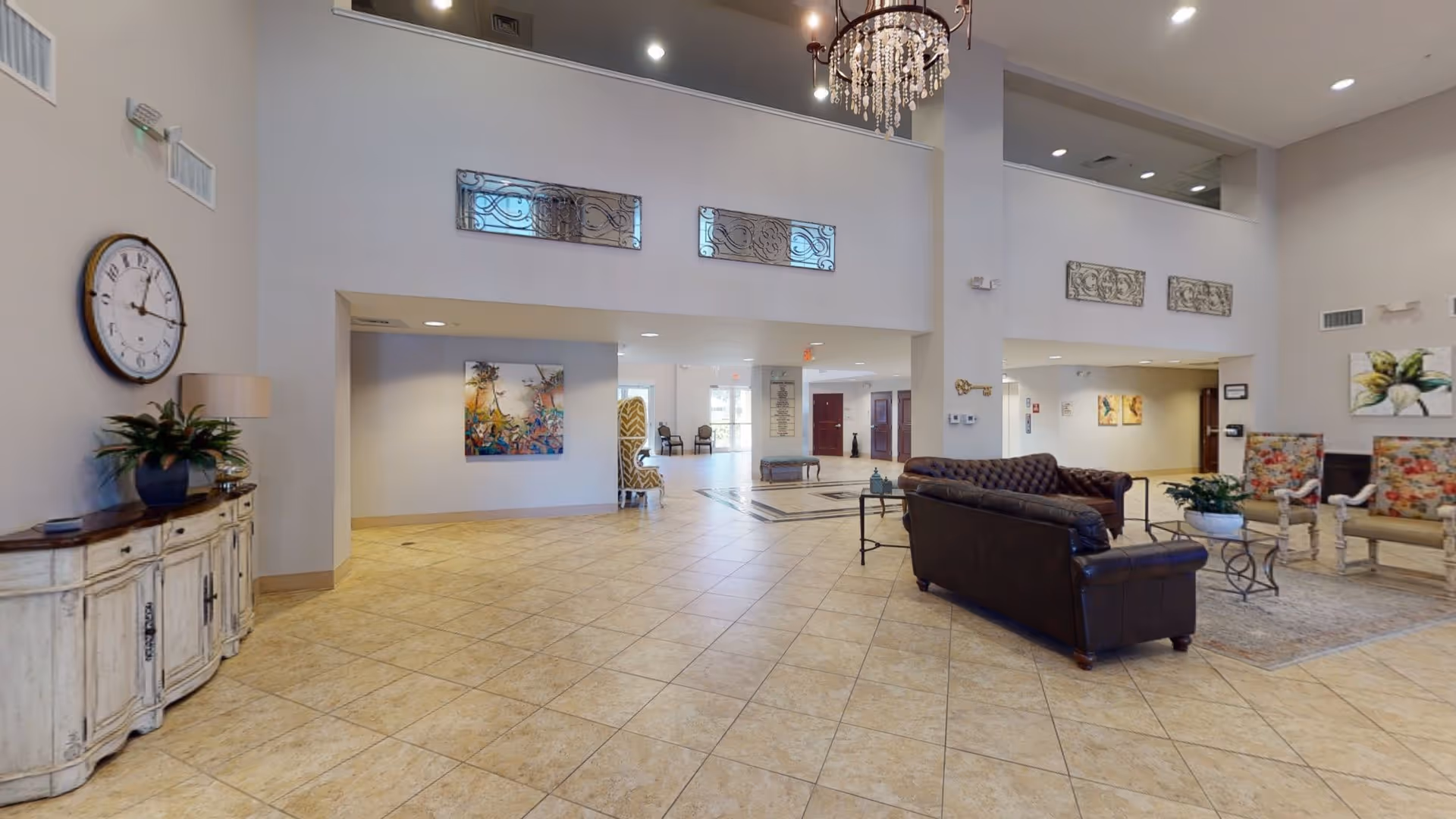 Spacious senior living facility lobby with beige tiled floor, high ceilings, and neutral-colored walls. The area features a large wall clock above a distressed white wooden console table with a plant and lamp. There are leather sofas and floral upholstered chairs arranged around a glass coffee table on a rug. Decorative wall art and a chandelier add to the welcoming atmosphere.