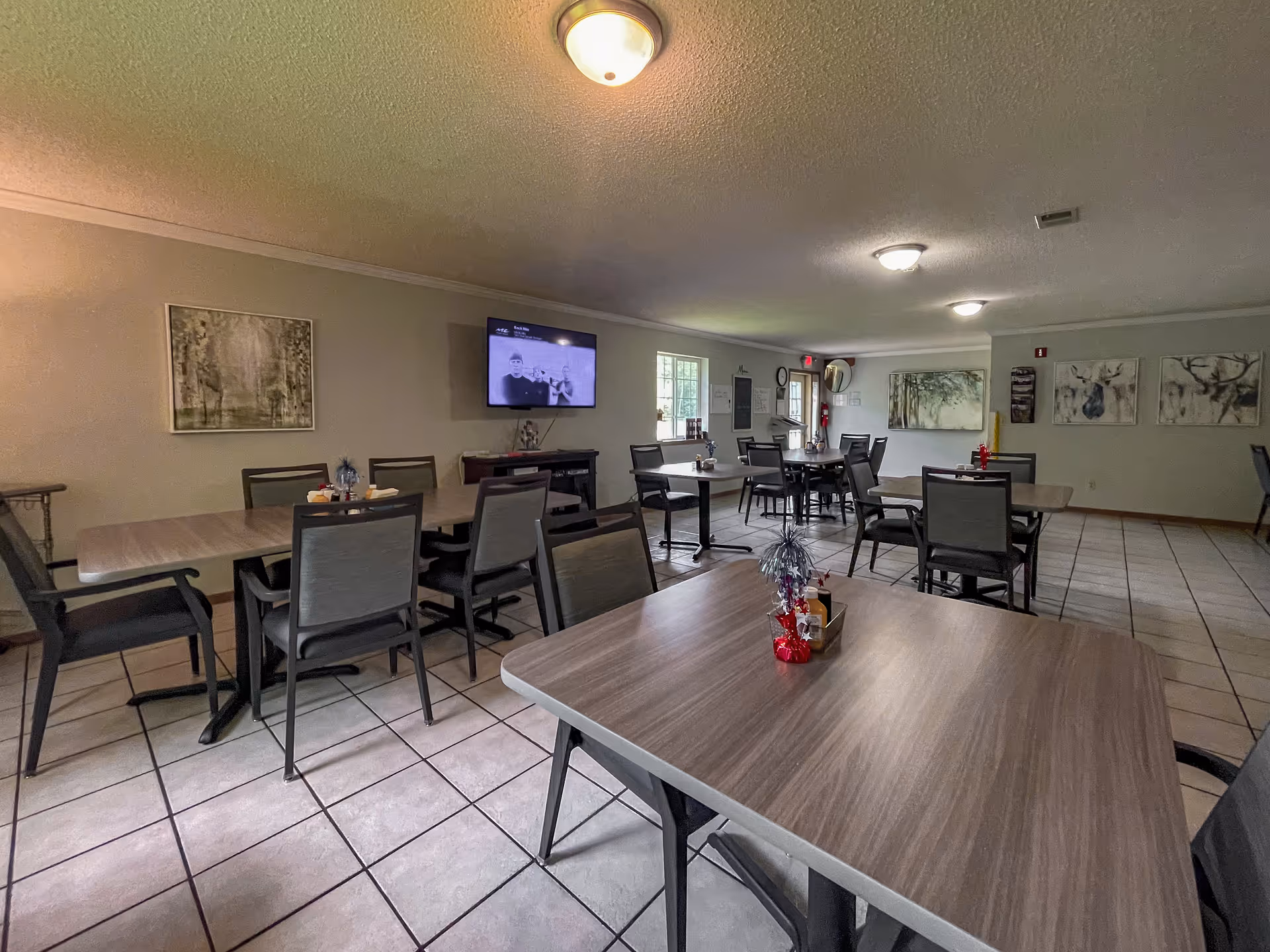 A spacious dining room with several tables and chairs arranged neatly. The room has tiled floors, light-colored walls adorned with artwork, and ceiling lights. A television is mounted on one wall, and windows allow natural light to enter the room.