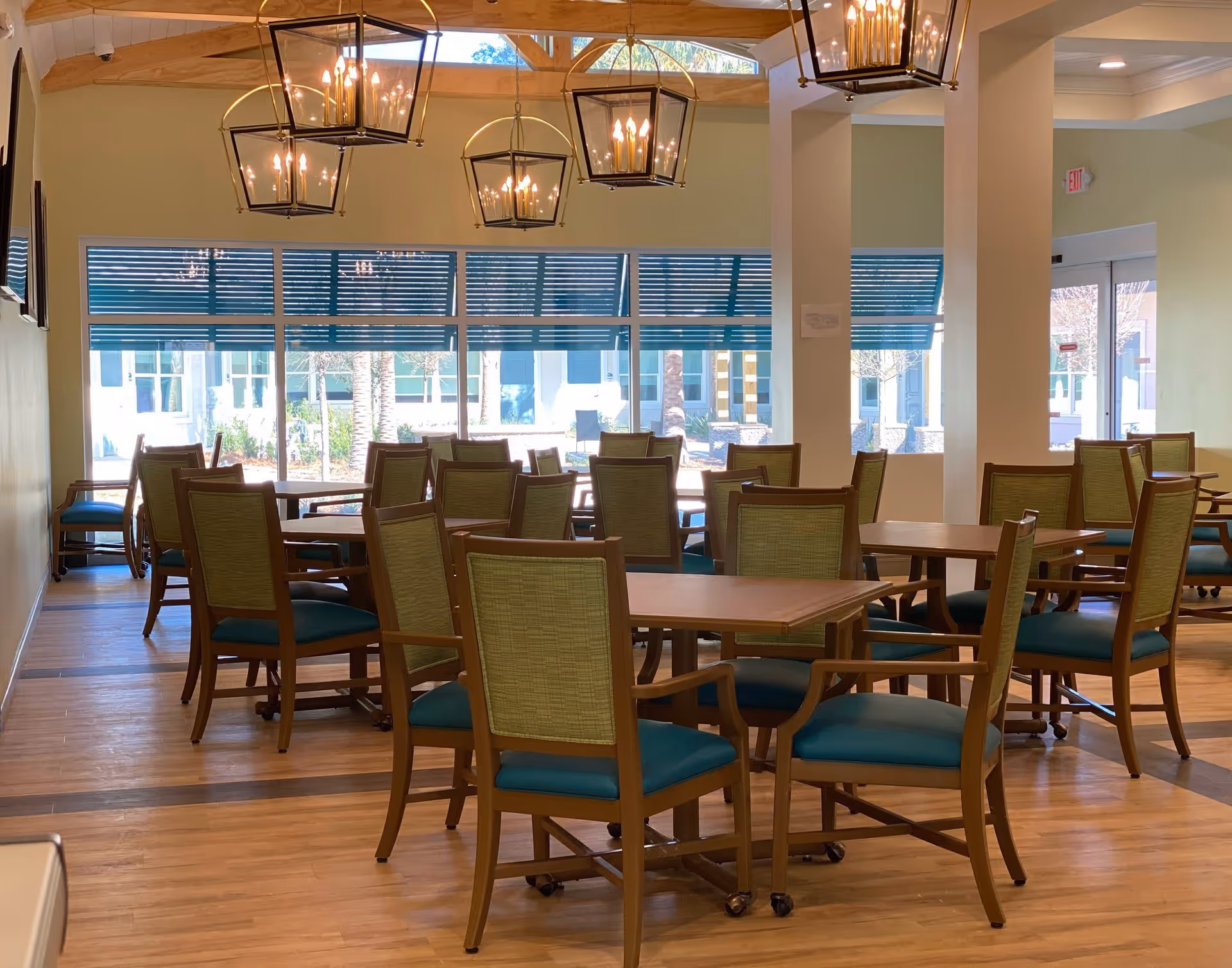 A bright dining room with multiple wooden tables and green cushioned chairs arranged neatly. Large windows with blue blinds allow natural light to fill the room. Several modern hanging light fixtures with multiple bulbs are suspended from the ceiling. The floor is wooden, and there is a glass door leading outside.