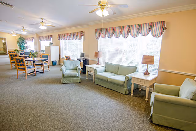 A spacious living room area in a senior living facility with light green upholstered sofas and armchairs, two white side tables with lamps, large windows with striped valances, ceiling fans, and a dining area with wooden chairs and tables in the background.