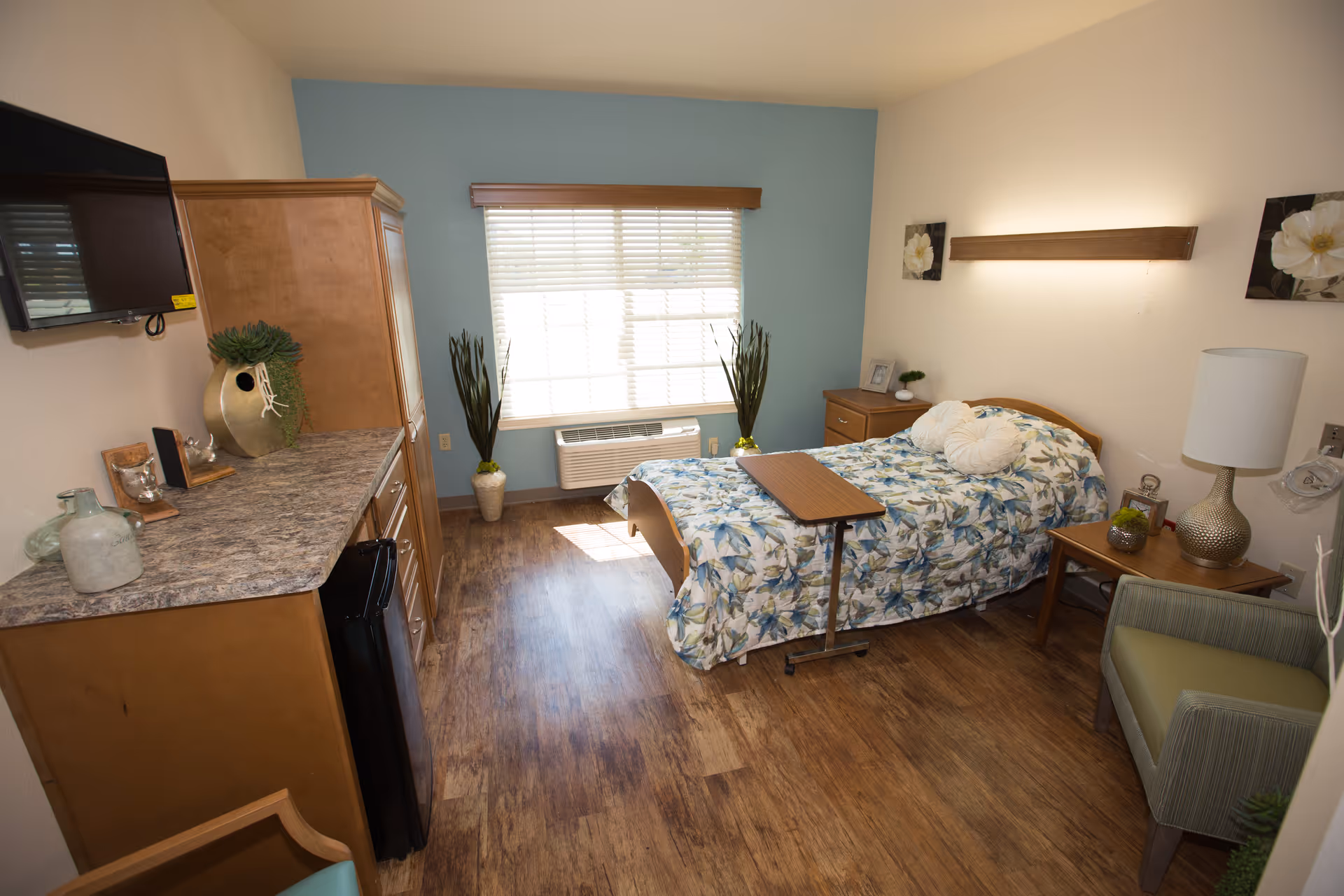 A well-lit bedroom in a senior living facility featuring a single bed with floral bedding and decorative pillows. The room includes a wooden nightstand with a lamp and small decor items, a green armchair, a wooden cabinet with a countertop holding decorative vases, and a wall-mounted TV. A large window with blinds allows natural light to fill the room, and the floor is covered with wood-style flooring.