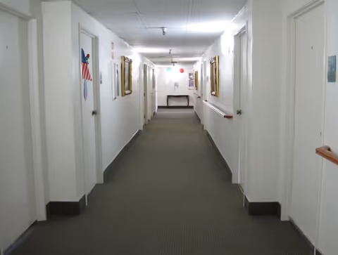 A long, narrow hallway in a retirement center with white walls and multiple closed white doors on both sides. The floor is carpeted in gray, and there are handrails along the walls. The ceiling has fluorescent lighting, and framed pictures hang on the walls. At the end of the hallway, there is a small table beneath a bulletin board.