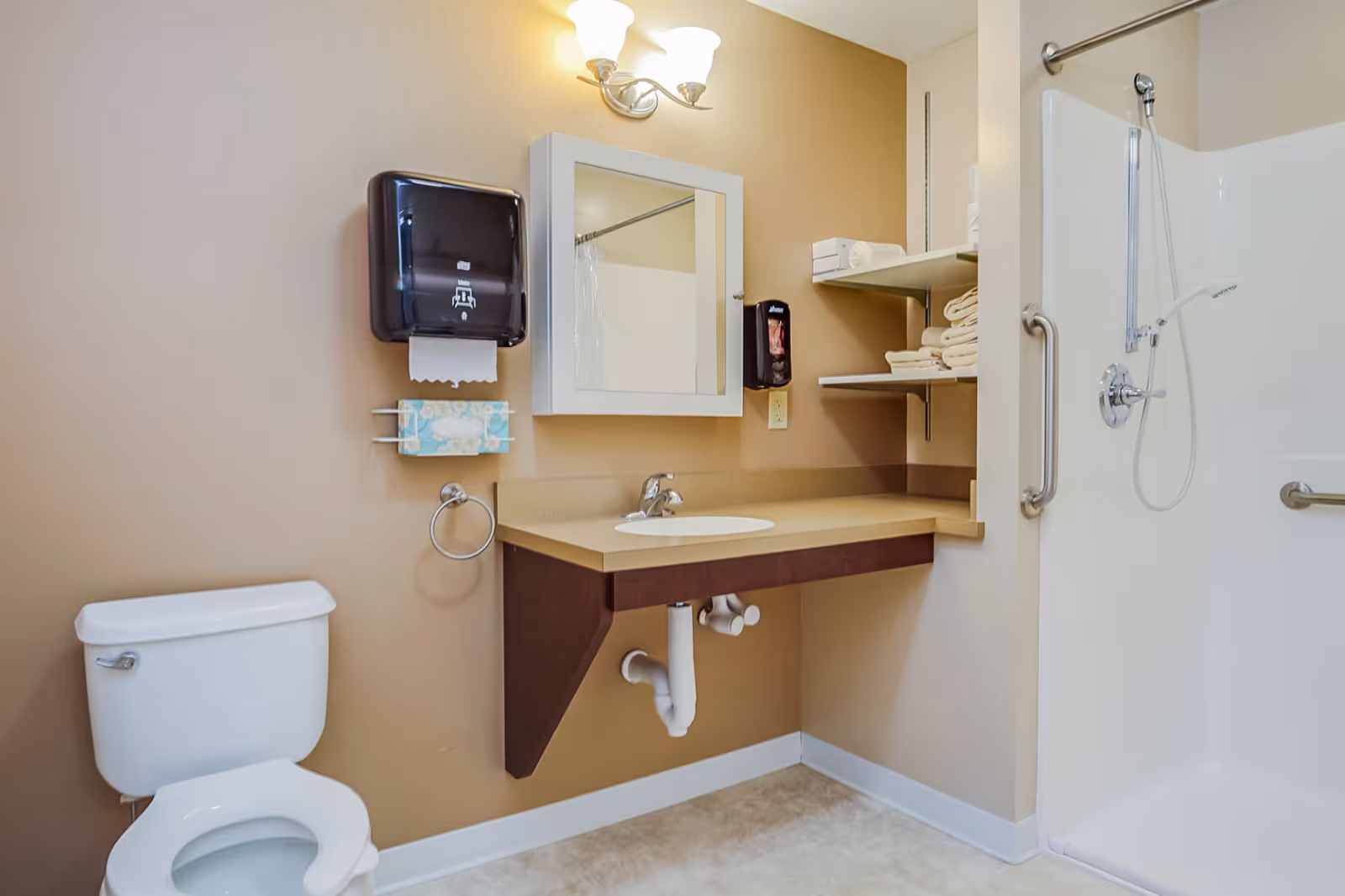 Accessible bathroom with a toilet, wall-mounted sink and mirror, paper dispenser and towel shelves, and a walk-in shower with grab bars.