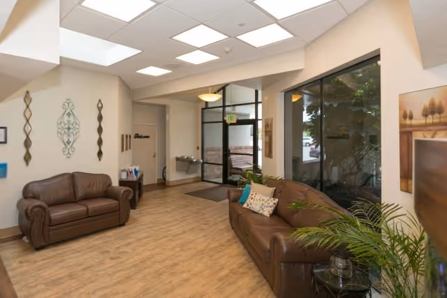 Lobby seating area with two brown leather sofas, wood-look flooring, large windows, plants and a glass entrance door.