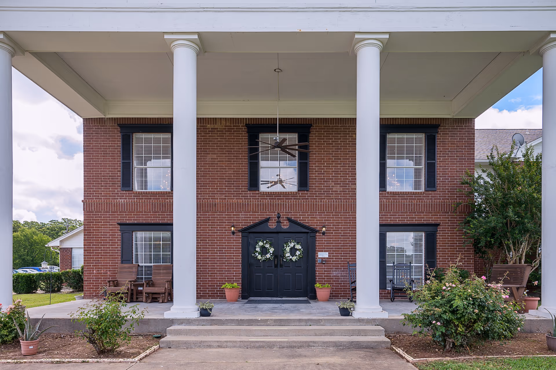 Front entrance of a brick building with four large white columns, black double doors decorated with wreaths, potted plants on either side of the entrance, and windows with black shutters. There are outdoor chairs and greenery around the entrance.