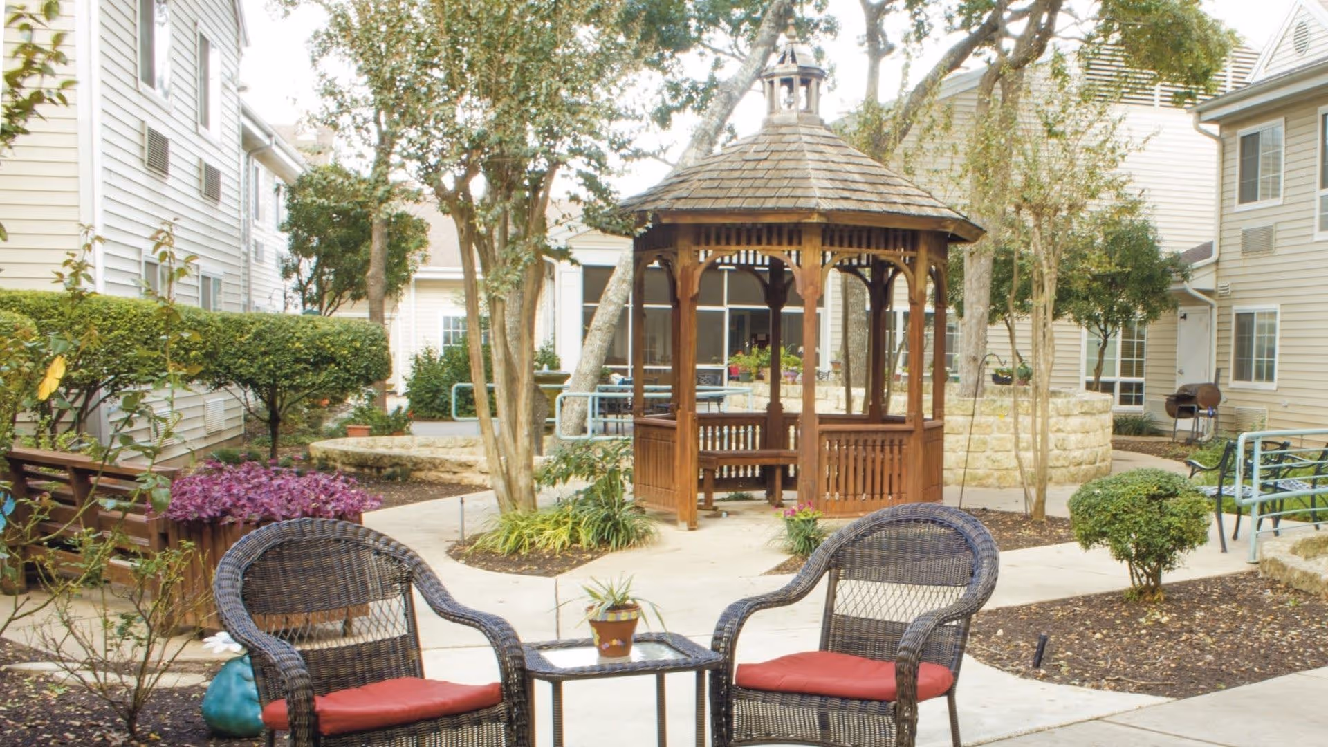 Outdoor courtyard area at Juniper Village at Spicewood Summit featuring a wooden gazebo, two wicker chairs with red cushions, a small table with a potted plant, surrounded by trees, shrubs, and residential buildings.