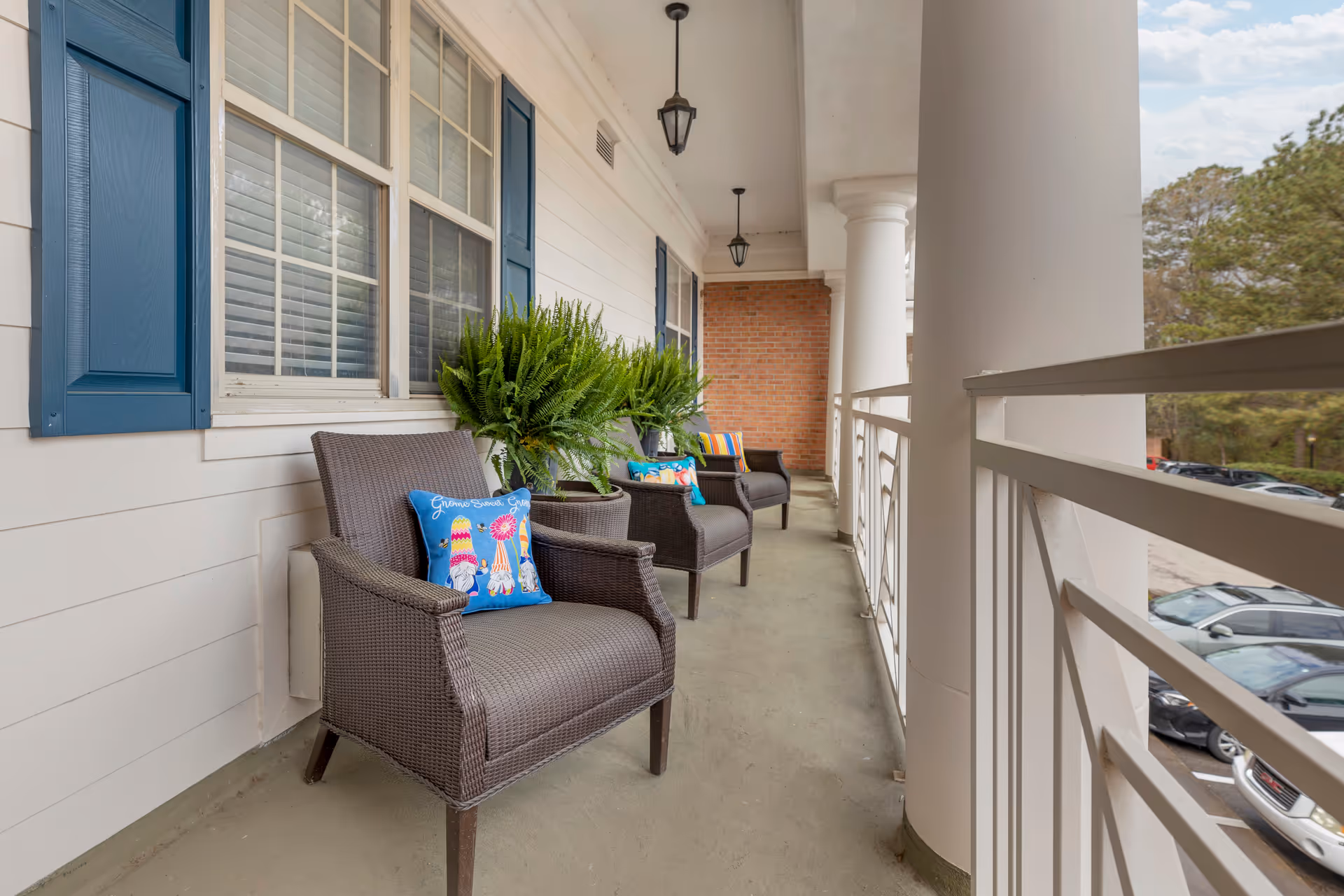 Covered balcony with wicker chairs, potted ferns and colorful pillows along a railing overlooking a parking lot.