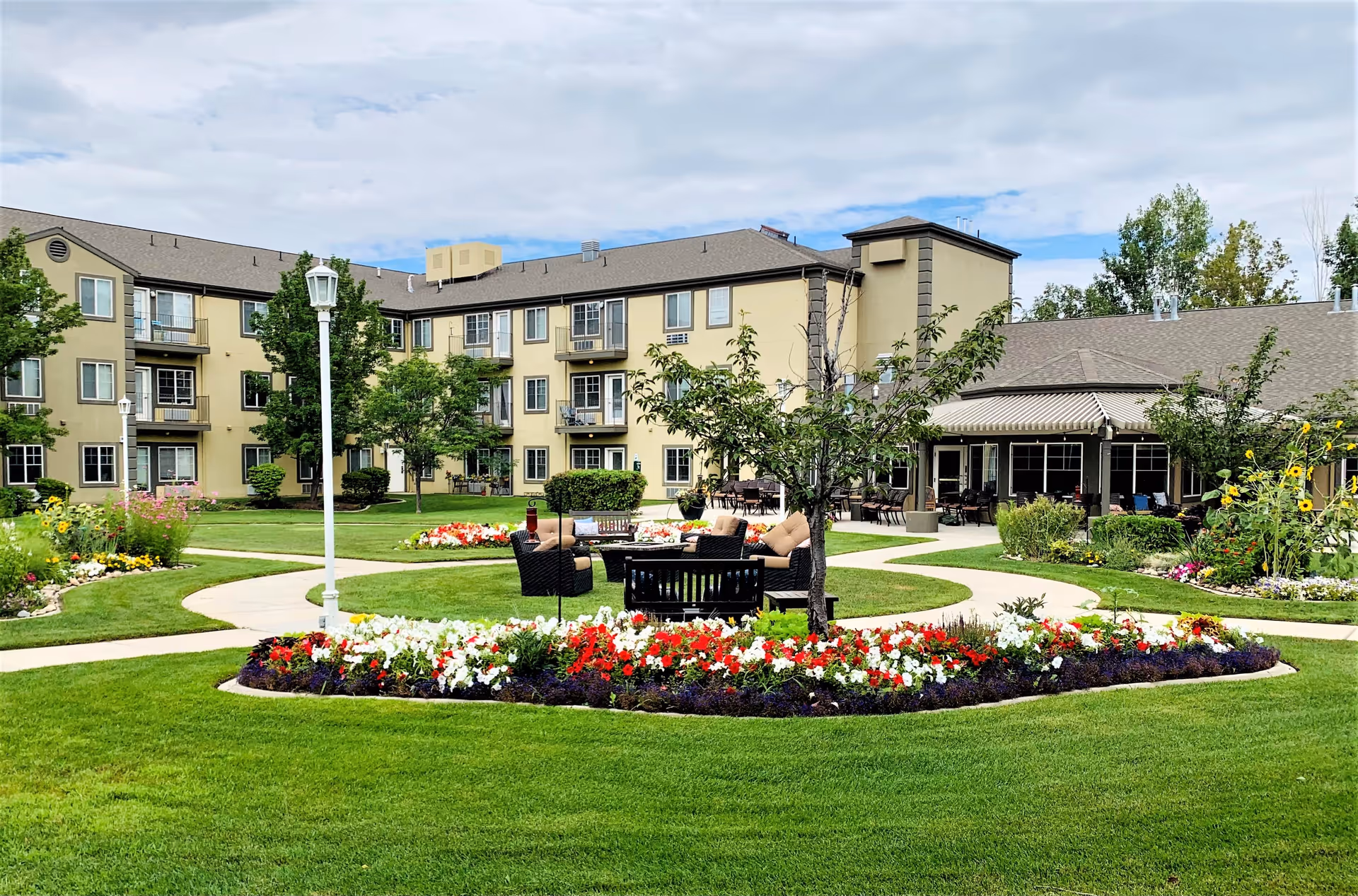 Outdoor garden area at Cedarwood at Sandy featuring a well-maintained lawn, colorful flower beds, paved walking paths, outdoor seating with cushioned chairs and a table, surrounded by a multi-story residential building under a partly cloudy sky.