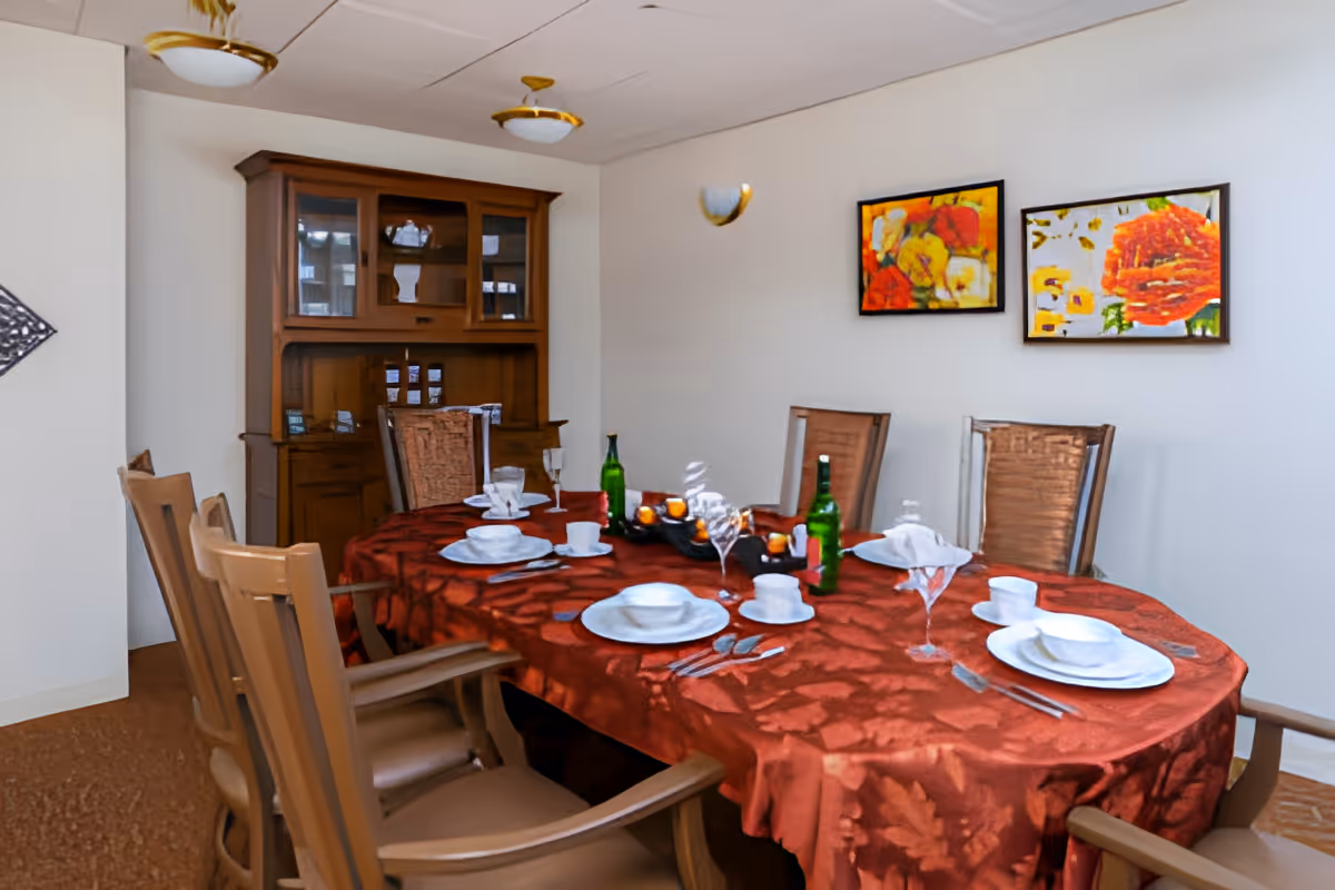 A dining room with a rectangular table covered with a red patterned tablecloth set with white plates, cups, silverware, and wine glasses. There are six wooden chairs around the table. A wooden cabinet with glass doors is against the wall, and two colorful floral paintings hang on the white wall above the table.