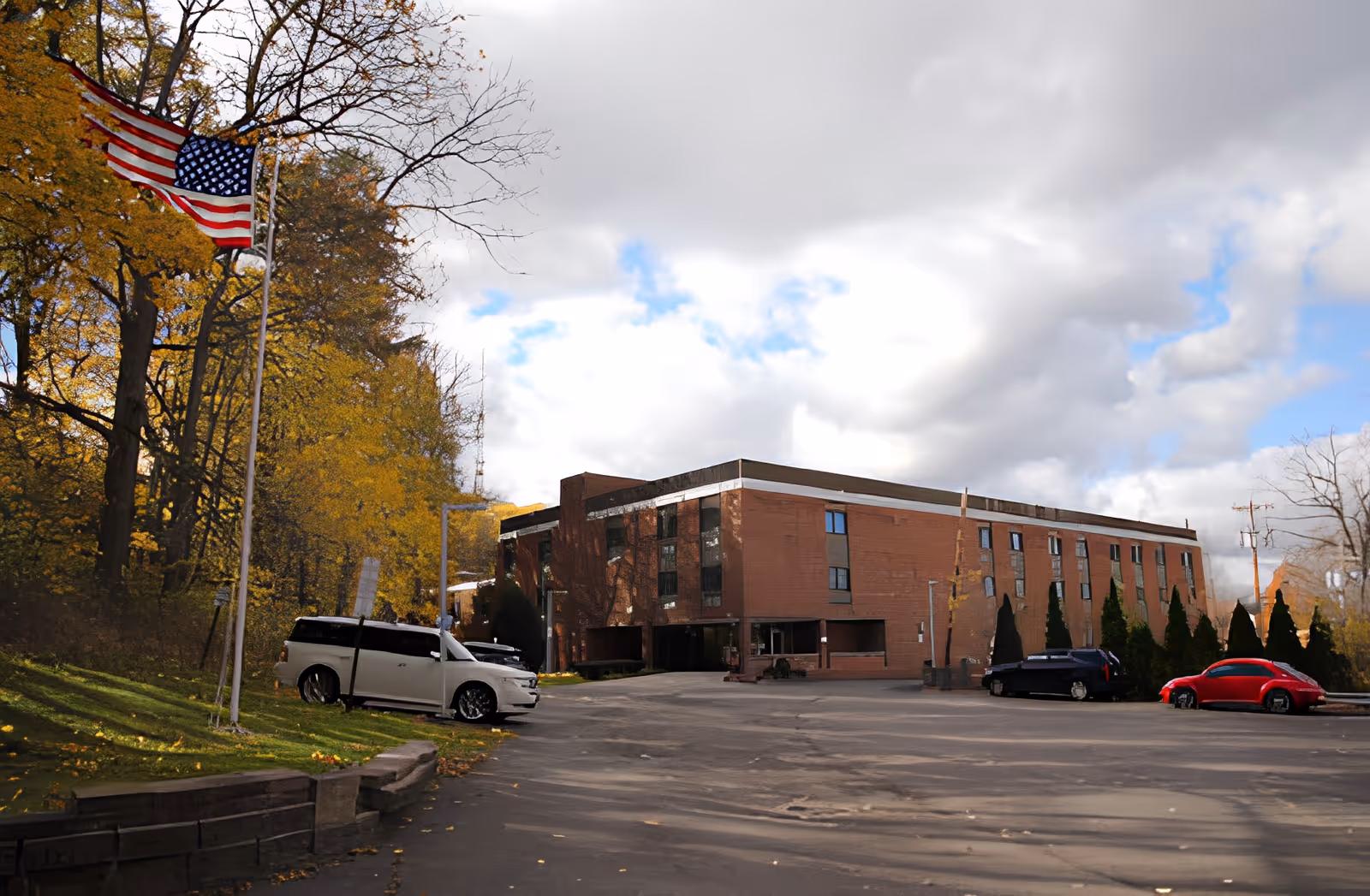 Exterior view of a three-story brick building with several windows, surrounded by trees with autumn foliage. There is an American flag on a flagpole to the left, and a few parked cars in the parking lot in front of the building under a partly cloudy sky.