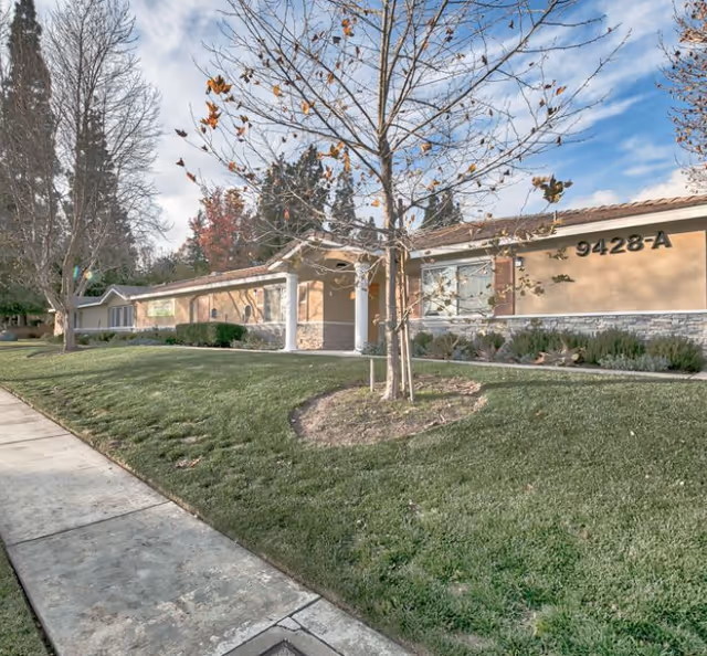 Exterior view of a single-story senior living facility with manicured lawn and sidewalk.