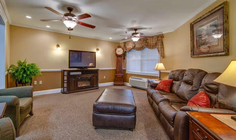 Cozy living room with a leather sofa, ottoman, TV above a fireplace, grandfather clock, and ceiling fans.