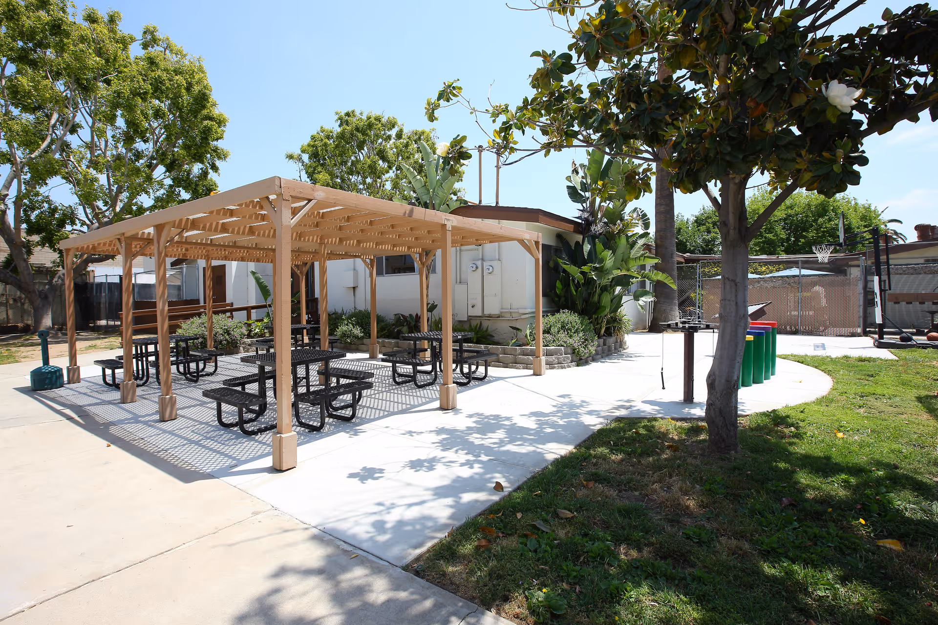 Outdoor seating area with multiple picnic tables under a wooden pergola surrounded by trees and greenery. There is a concrete pathway and a grassy area nearby with a basketball hoop and recycling bins in the background.