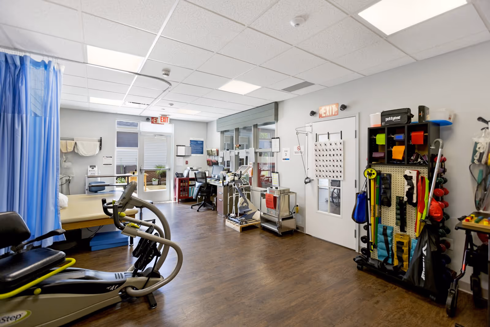 A physical therapy or rehabilitation room with exercise equipment including a recumbent stepper, therapy bed, and various therapy tools organized on a rack. There is a desk with a computer and office supplies near the back, and two exit signs above doors. The room has wood flooring and a drop ceiling with fluorescent lighting.