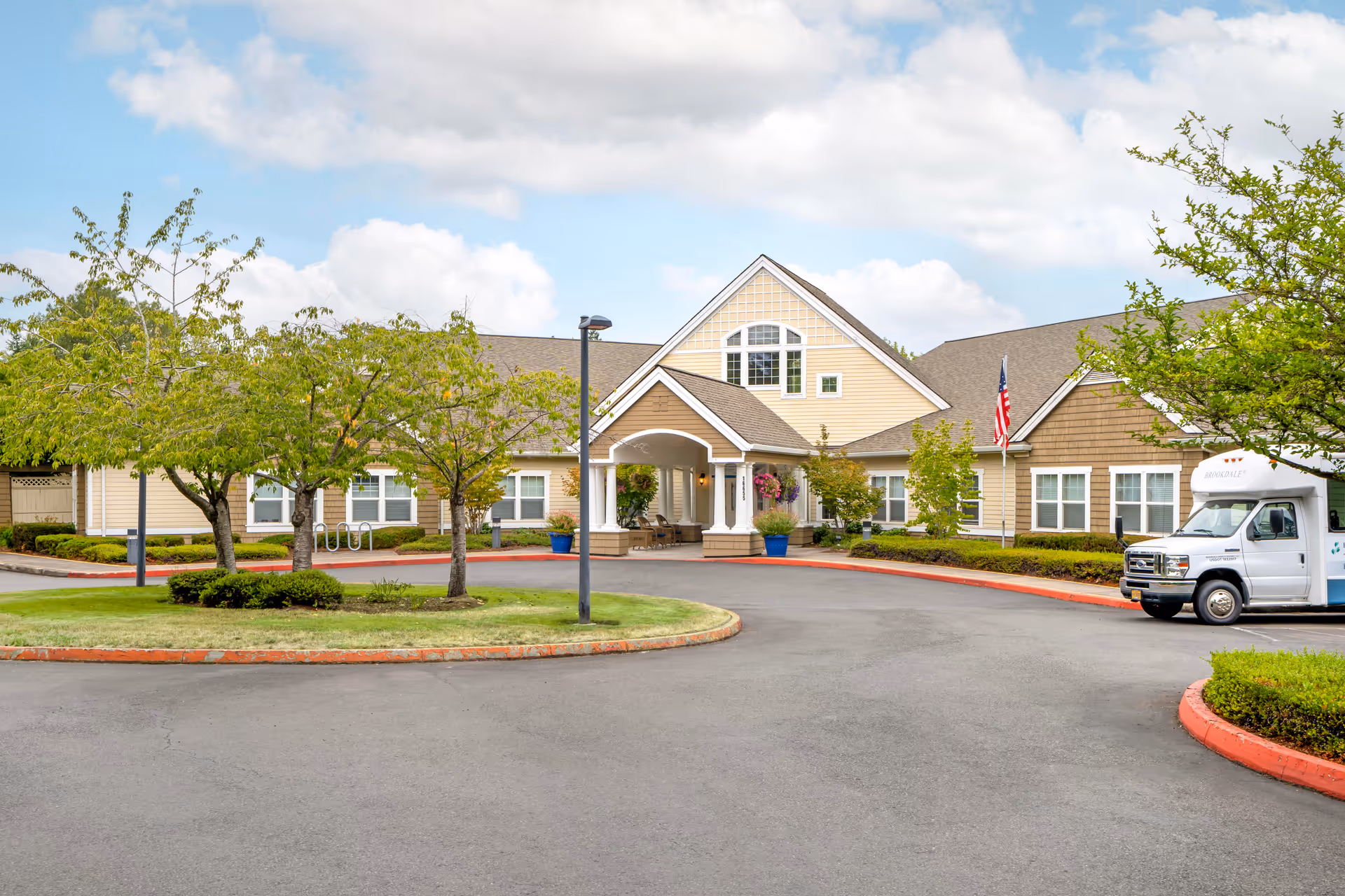 Exterior view of Brookdale Beaverton senior living facility showing a single-story building with a covered entrance, surrounded by trees and landscaping under a partly cloudy sky. A white shuttle van is parked near the entrance.