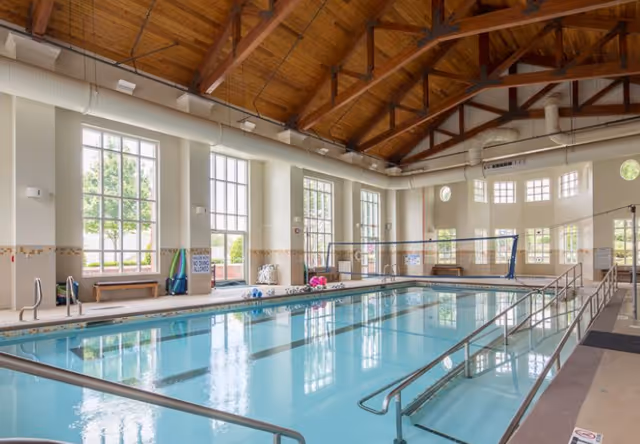Indoor swimming pool area with clear blue water, metal handrails, and a volleyball net across the pool. The room has large windows letting in natural light, wooden ceiling beams, and benches along the walls.