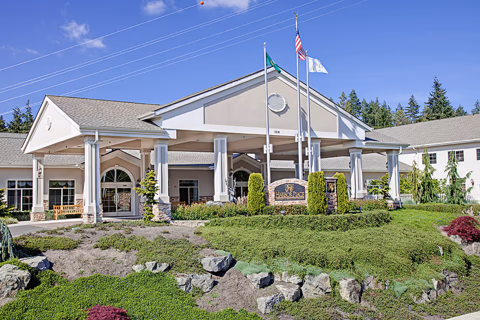 Front exterior view of Harbor Place at Cottesmore, a senior living facility with a covered entrance, three flagpoles with flags, landscaped greenery, and a clear blue sky.