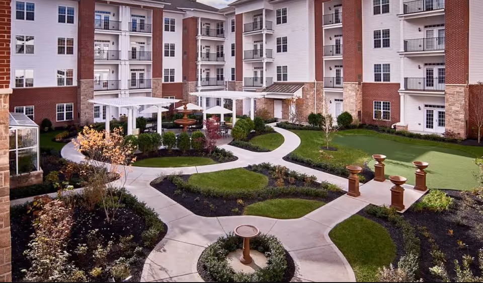Landscaped courtyard with winding walkways, pergolas, a fountain, and surrounding multi-story senior living building balconies.