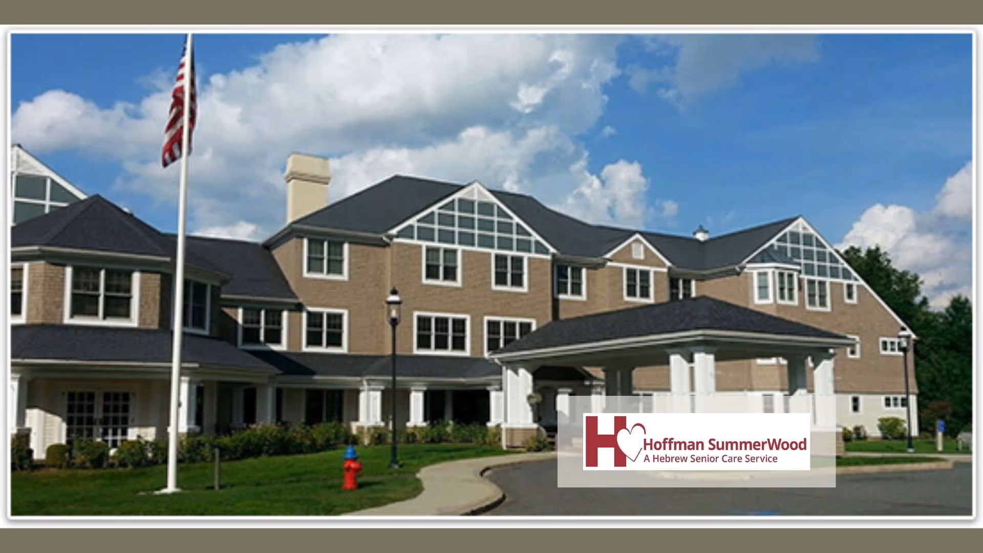Exterior view of a large senior living facility building with multiple windows, a covered entrance, an American flag on a flagpole, and a well-maintained lawn under a partly cloudy sky.