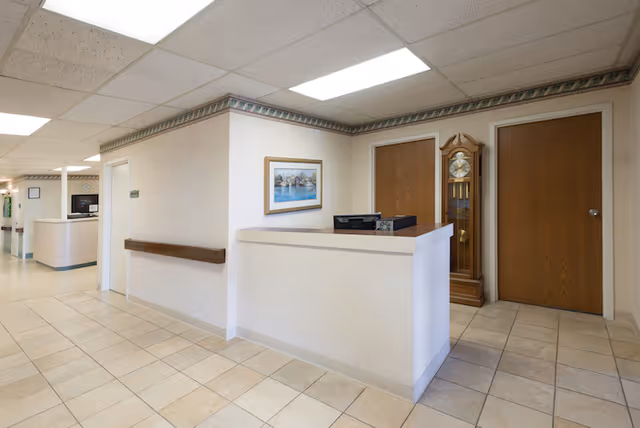 Reception desk and hallway inside a senior living facility with tile floors, a grandfather clock, framed art, and two wooden doors.
