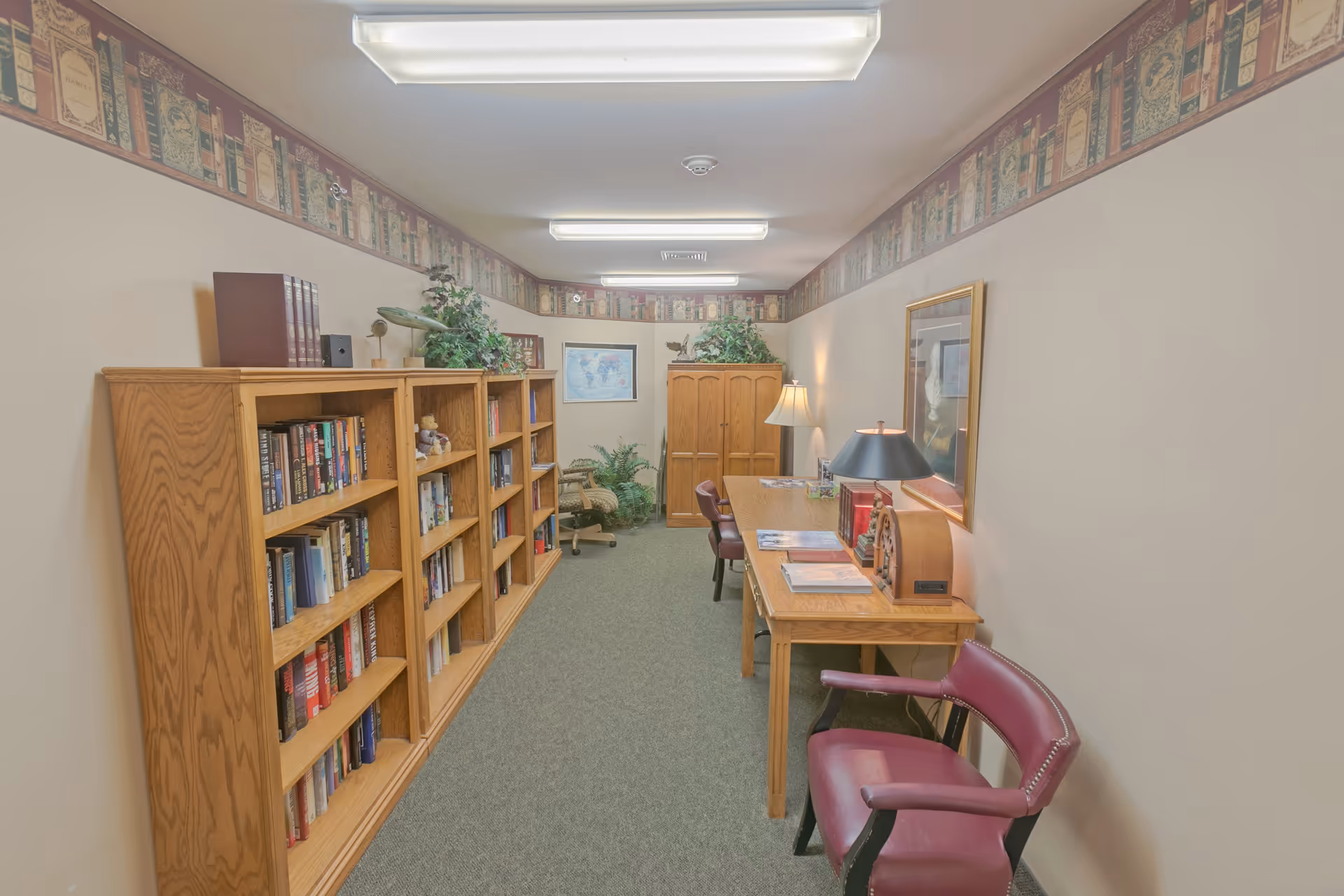 A narrow room with wooden bookshelves filled with books on the left side and a wooden table with chairs on the right side. The room has beige walls with a decorative border near the ceiling, carpeted floor, and fluorescent ceiling lights. There are plants and framed pictures on the walls, creating a cozy reading or study area.