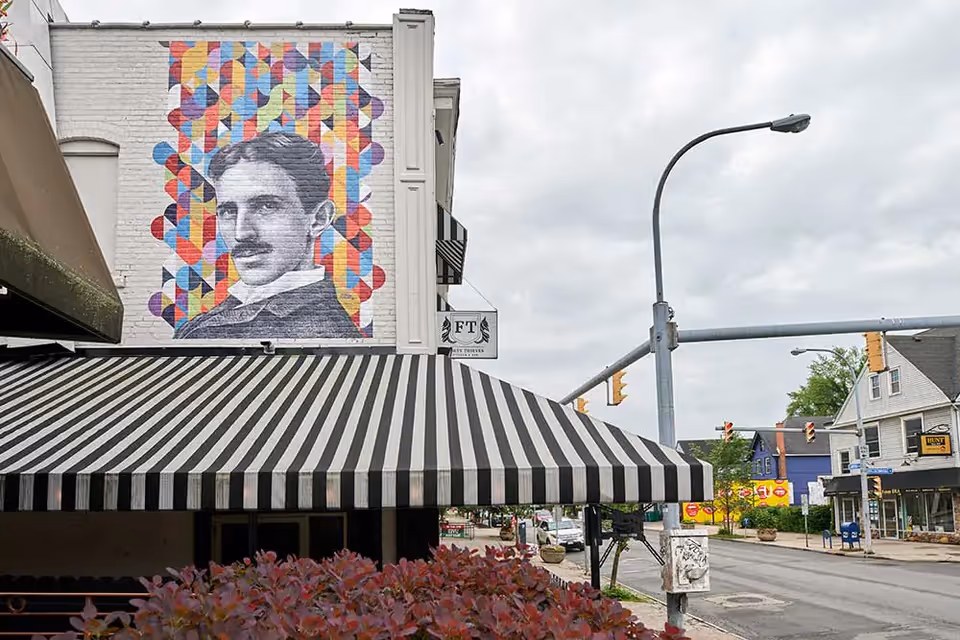 Street view showing a building with a large mural of a man with a mustache on a colorful geometric background. The building has a black and white striped awning over the entrance. Traffic lights and other buildings are visible in the background under a cloudy sky.