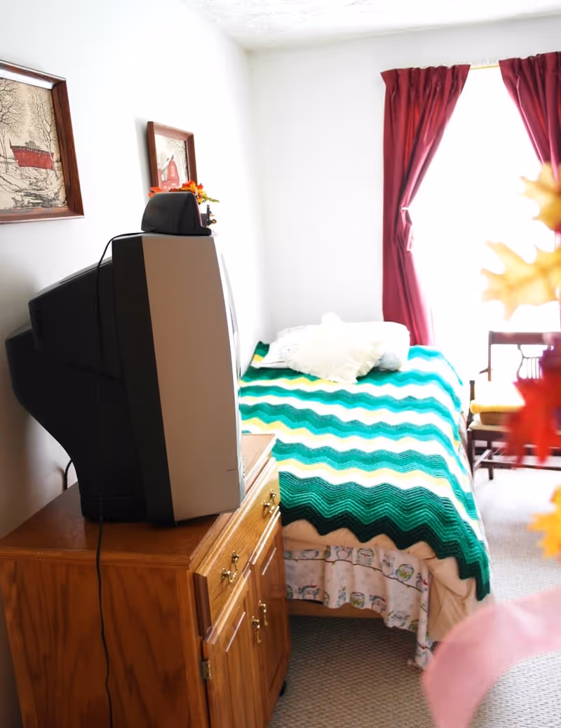 A small bedroom with a single bed covered by a green, yellow, and white zigzag patterned blanket. There is a wooden cabinet with an old-style CRT television on top. The room has white walls with two framed pictures hanging, a window with red curtains, and a wooden chair with a yellow cushion near the window.