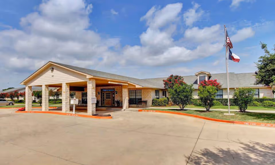 Exterior view of Sagebrook Nursing and Rehabilitation facility showing a single-story building with a covered entrance supported by stone pillars, a driveway, landscaped bushes, and two flagpoles with the American and Texas flags against a partly cloudy sky.