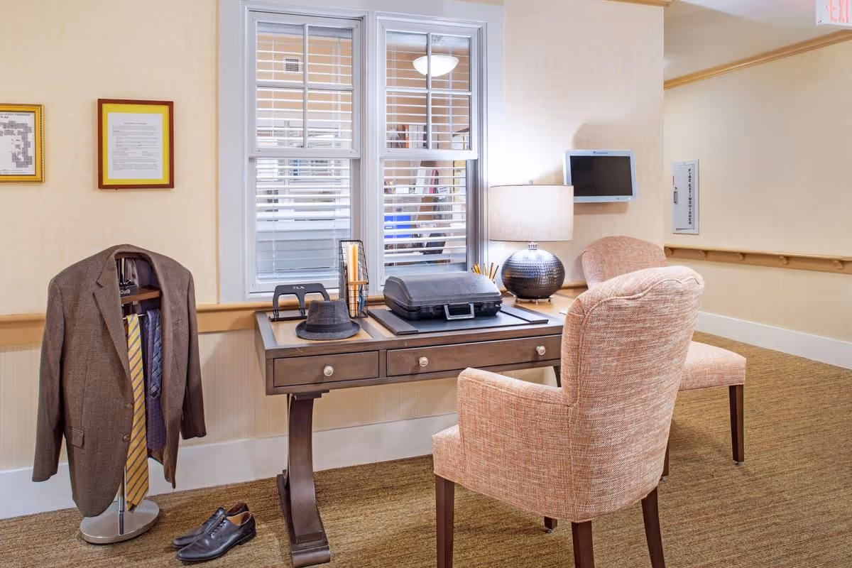 A small office or workspace area in a senior living facility with a wooden desk, two upholstered chairs, a lamp, and office supplies. A suit jacket, ties, and shoes are displayed on a stand next to the desk. There is a window with white blinds behind the desk and a small wall-mounted TV on the right side.