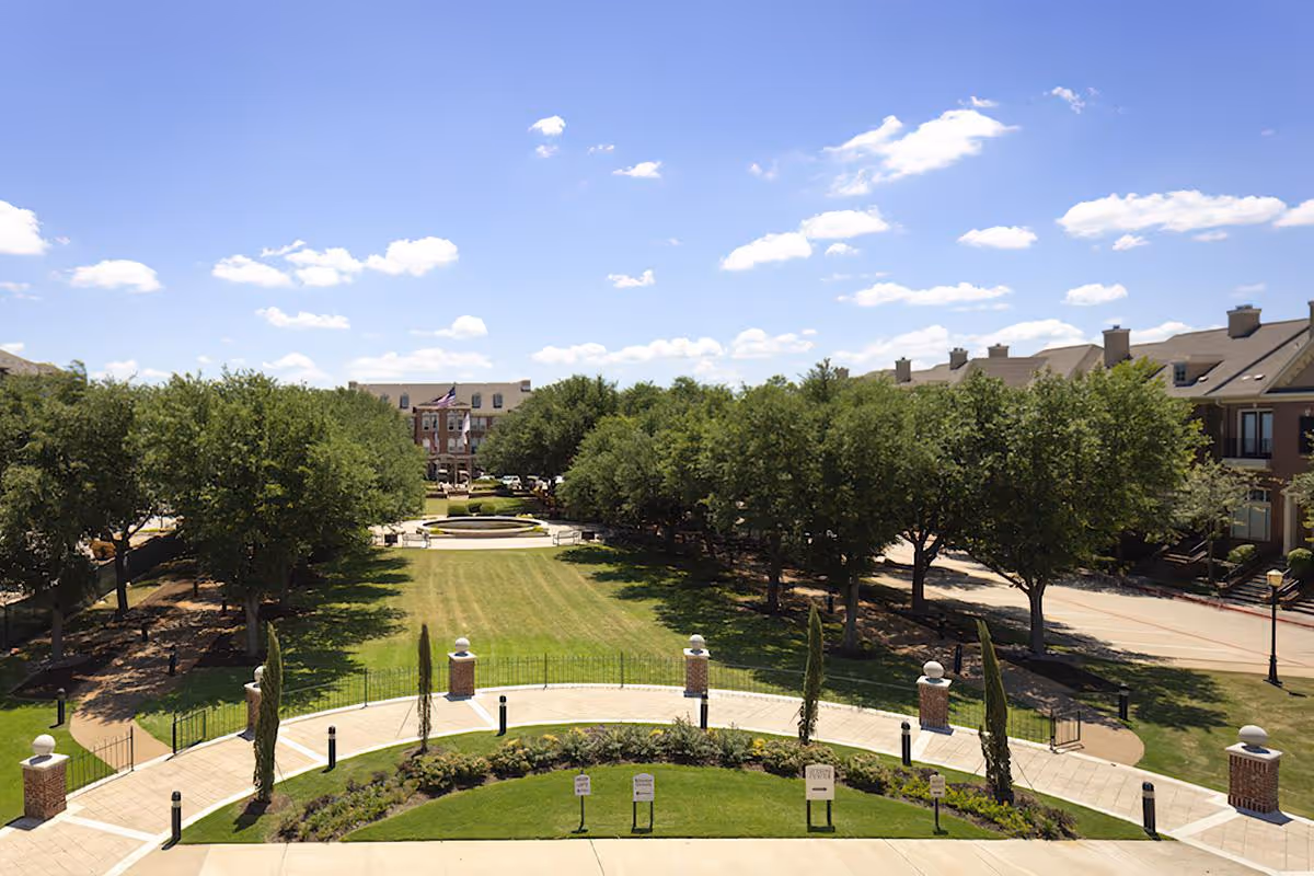A sunny outdoor view of McDermott Crossing featuring a wide green lawn bordered by trees on both sides, a paved walkway with brick pillars and small shrubs, and residential buildings in the background under a blue sky with scattered clouds.
