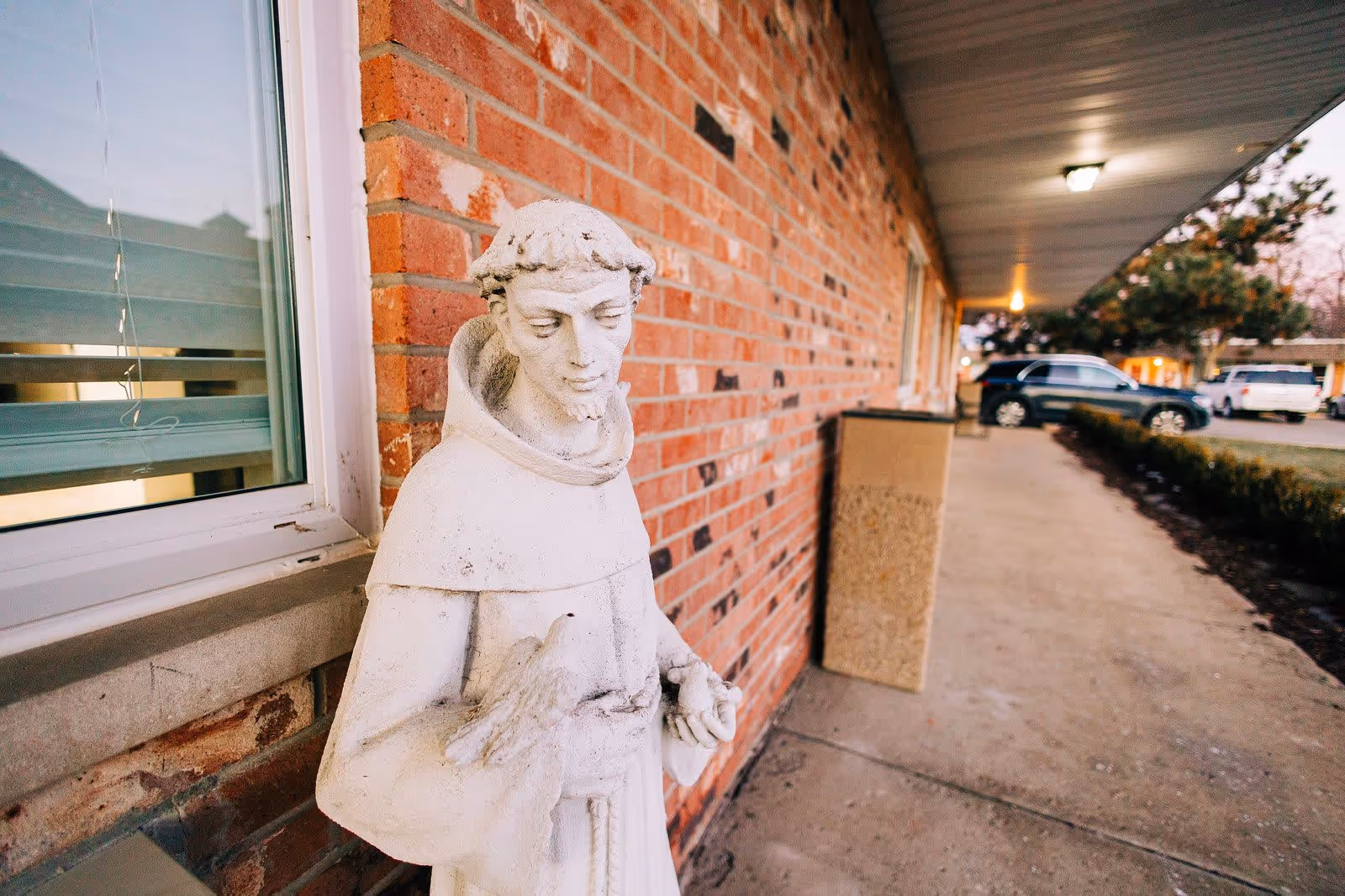 A white statue of a robed figure holding a bird, positioned outside next to a brick wall and a window. The scene includes a covered walkway with parked cars and trees in the background.