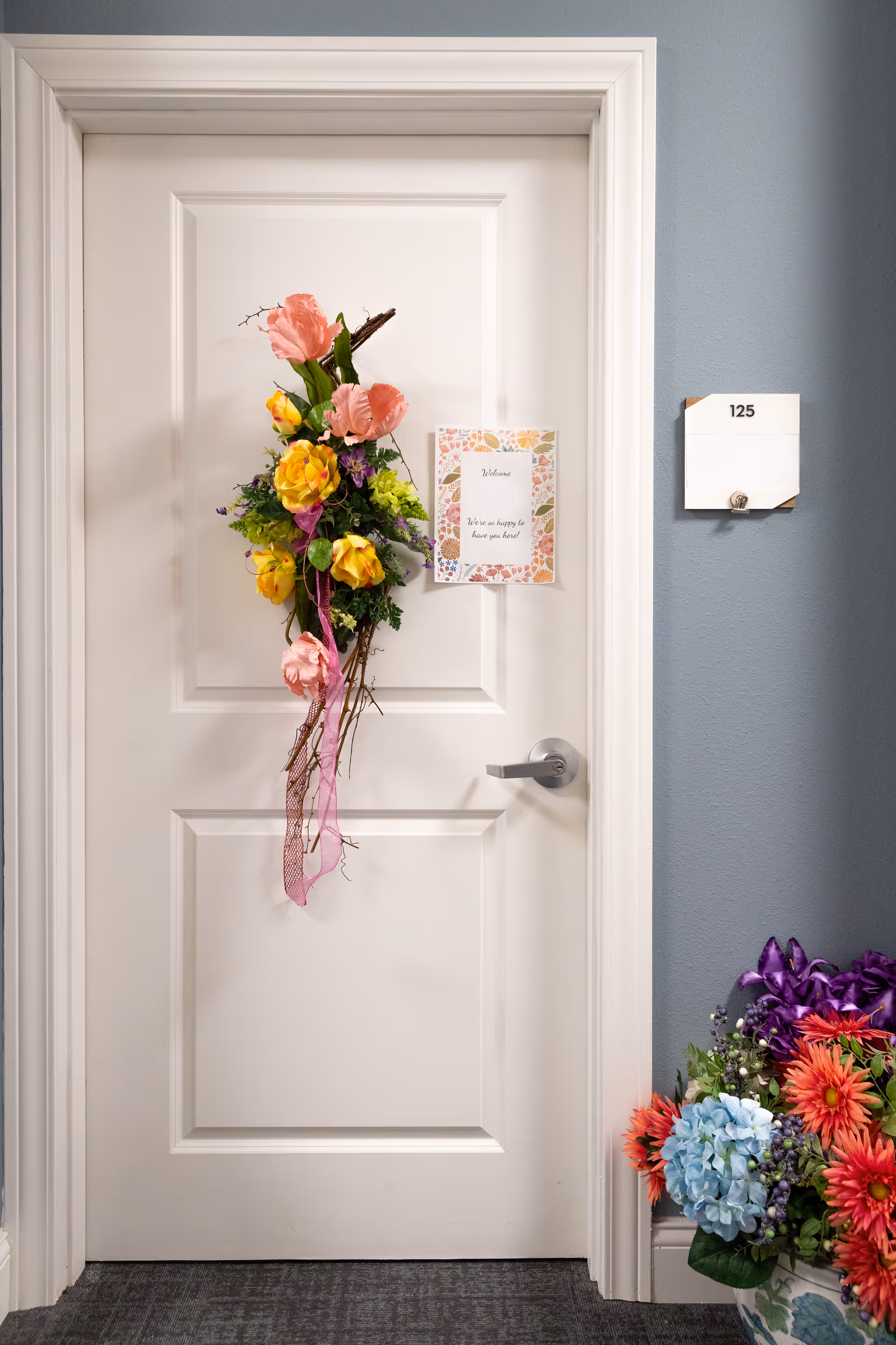 White interior door decorated with a hanging floral arrangement and welcome card, room number 125 on the wall and a bouquet at the base.