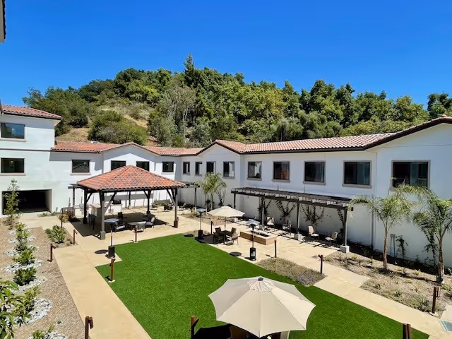 Outdoor courtyard area of Tuscan Hills Senior Living with a green lawn, several patio umbrellas, seating areas, and a covered gazebo. The building surrounding the courtyard is white with a red-tiled roof, and there are trees and hills in the background under a clear blue sky.