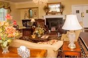 A cozy living room in Bethea Retirement Community featuring a beige sofa with floral cushions, a wooden coffee table with a floral arrangement, a fireplace with decorative items on the mantel, a large mirror above the fireplace, an armchair, a side table with a lamp, and a chandelier hanging from the ceiling. The room has warm lighting and traditional decor.