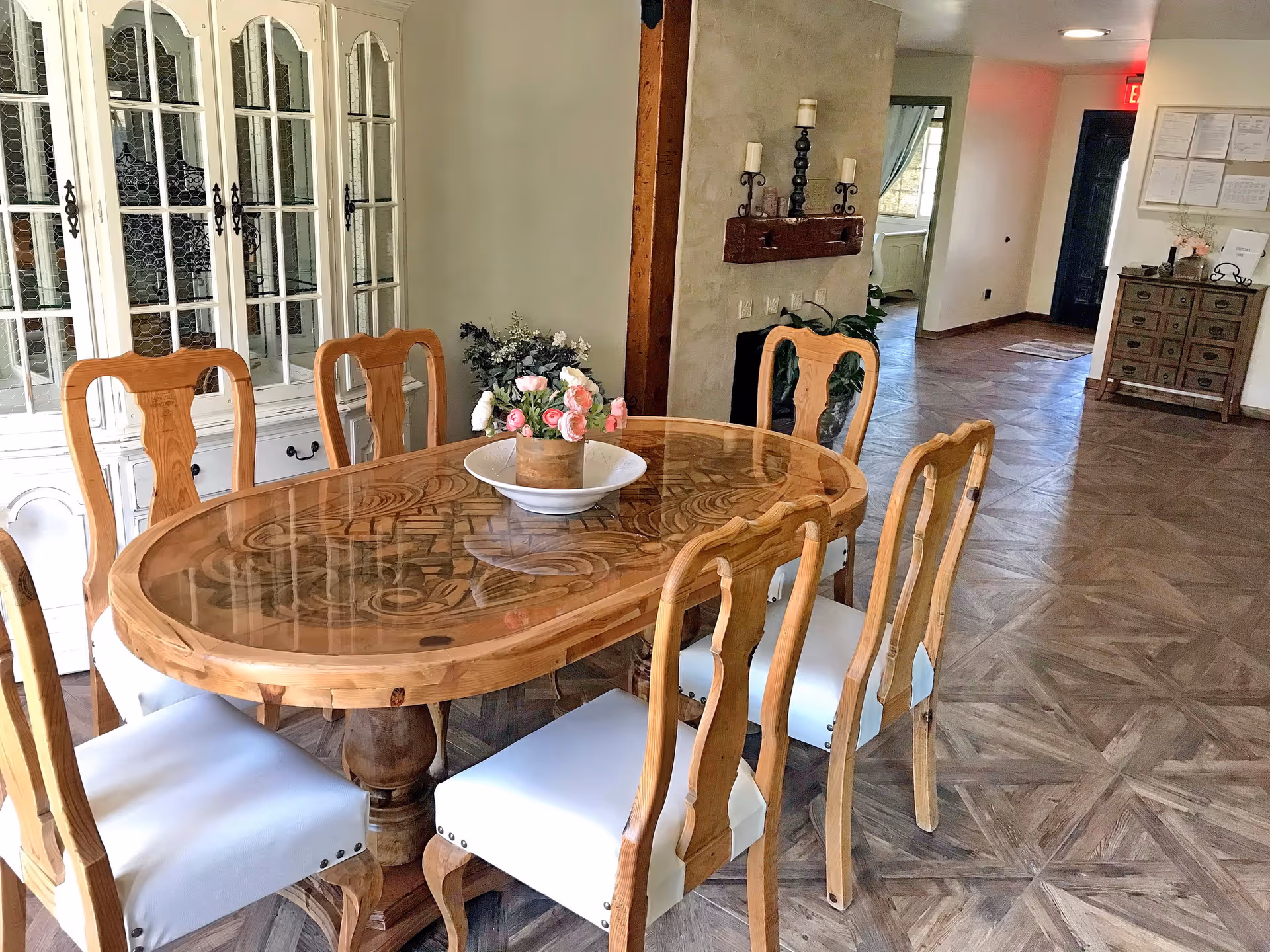 A dining area with a wooden oval table featuring intricate carvings and a glass top, surrounded by six wooden chairs with white cushioned seats. A flower arrangement in a vase sits in the center of the table. In the background, there is a white cabinet with glass doors, a wall with candle holders, and a hallway leading to other rooms.