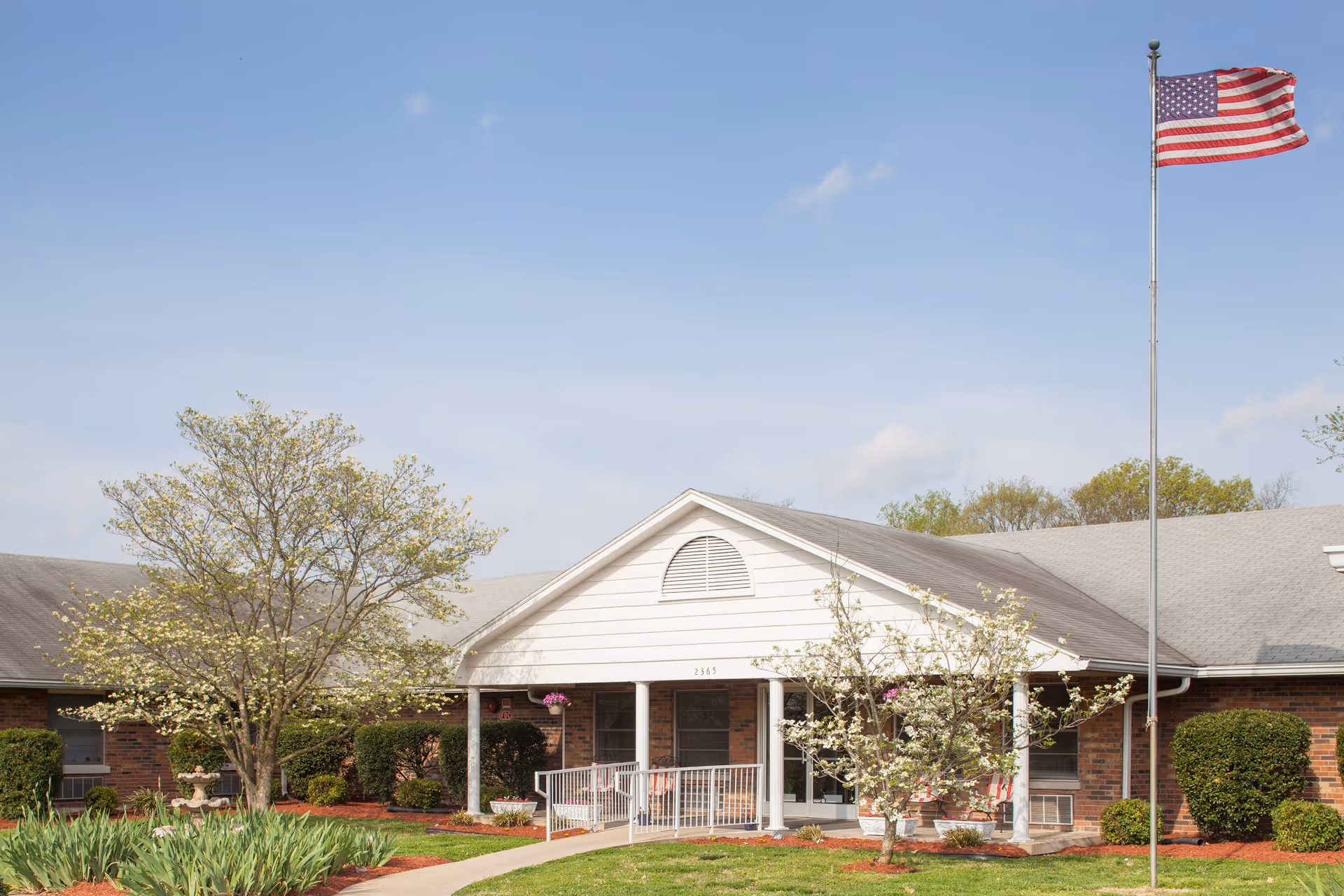 Exterior view of the Colonial Center building with a white entrance porch, surrounded by trees and bushes. An American flag is flying on a flagpole to the right of the entrance under a clear blue sky.