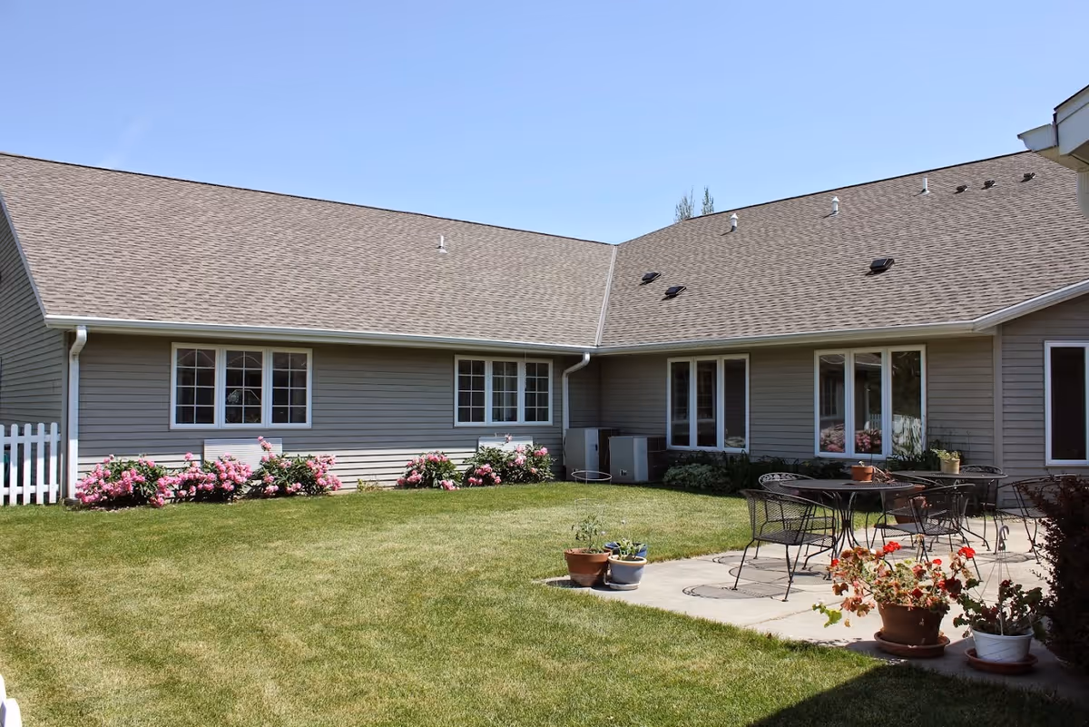 Outdoor patio area of a single-story building with beige siding and multiple windows. The patio has a metal table and chairs, surrounded by potted plants and flowers. The lawn is green and well-maintained with flower beds along the building.