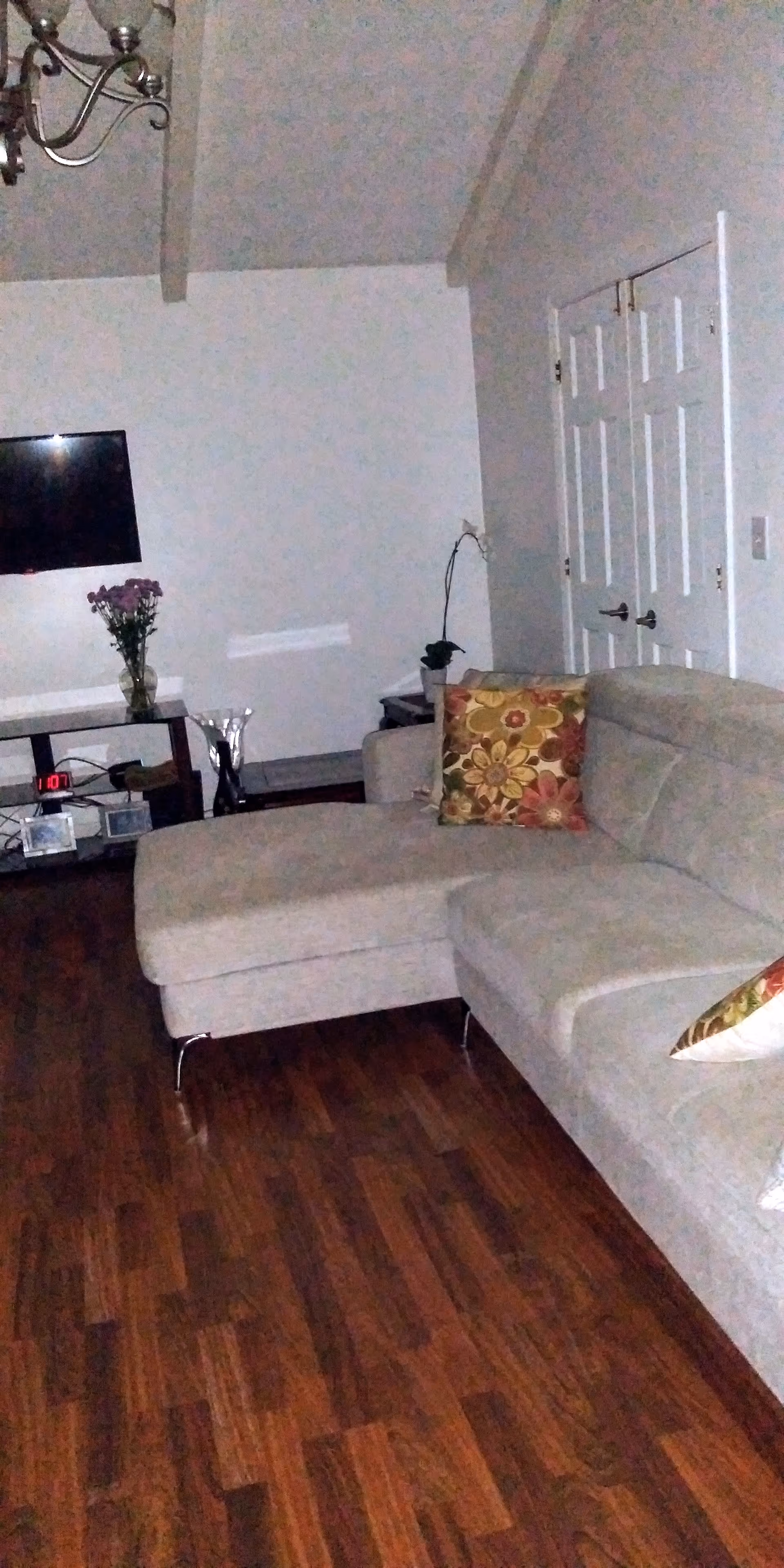 Interior view of a living room with a beige sectional sofa adorned with floral patterned pillows. The room has wooden flooring, a wall-mounted flat-screen TV, a small table with a vase of flowers, and double white doors. A chandelier is partially visible on the ceiling.
